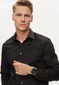 Chemise noire à boutons, au design épuré, avec un col et un détail de logo. Modèle portant une montre en argent avec un cadran noir.