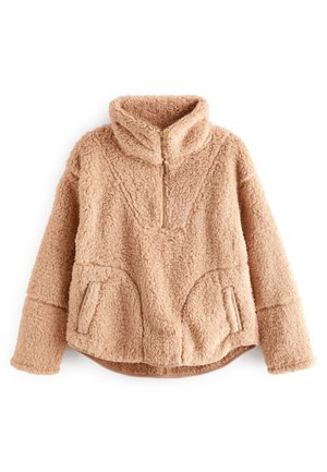 Personne portant une veste polaire beige, des leggings noirs, des chaussettes blanches et des bottines beige, debout devant un canapé avec des coussins et un tapis tissé.