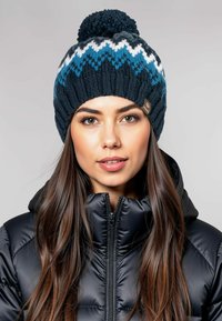 Gebreide beanie in donkerblauw met witte en turquoise zigzagpatronen, afgewerkt met een pompon. Voorzien van een merketiket aan de zijkant.