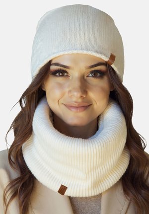 Kvinde iført en hvid strikket hue og en matchende chunky, ribstrikket halsvarmer, smilende blidt, med langt brunt hår synligt.