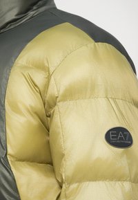 Rękaw kurtki puchowej w oliwkowym zielonym i ciemnoszarym kolorze z czarną, owalną łatą logo EA7 Emporio Armani na górnej części ramienia.