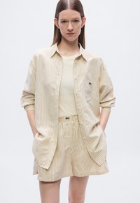 Μπεζ ριγέ πουκάμισο με κοντά μανίκια και τσέπη, σε συνδυασμό με σορτς με ελαστική μέση που ταιριάζουν. Και τα δύο έχουν μια μικρή διακοσμητική λεπτομέρεια με λογότυπο.