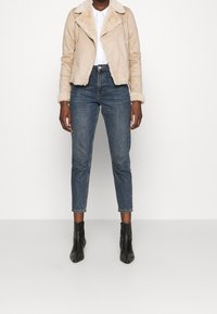 Chaqueta de motorista de ante beige con cuello de pelo sintético, llevada sobre una camisa blanca, combinada con unos vaqueros de mezclilla azul y botas negras de tobillo.