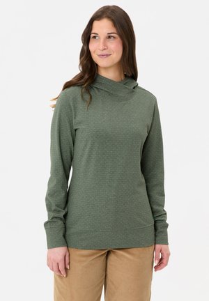 Jonge vrouw met lang bruin haar, gekleed in een groen shirt met lange mouwen en beige broek, staand tegen een effen witte achtergrond.