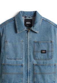 Lyseblå denimjakke med lynlåslukning, spids krave og to forlommer. Har et lille Vans-logo på venstre lomme.