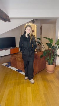 Juodas paltas su užtrauktuku, apykakle ir diržu, derinamas su juodomis kelnėmis ir šviesiais sportbačiais. Vidaus aplinka su medinėmis grindimis ir augalu.