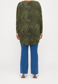 Grön leopardmönstrad långärmad tröja med avslappnad passform, kombinerad med blå utsvängda jeans. Tröjan har en rundad nederkant, vilket ger en avslappnad look.