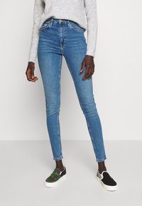 Skinny blue denim jeans with a high waist, featuring stitched pockets and a slight fade. Paired with patterned slip-on shoes.