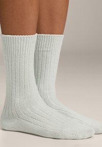 Un paio di piedi che indossano calze a coste grigio chiaro fino al polpaccio su uno sfondo beige uniforme.