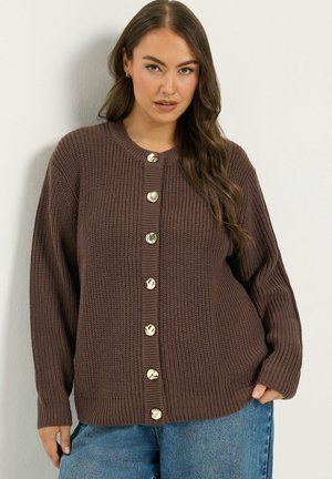 Donna che indossa un cardigan marrone di maglia con grandi bottoni rotondi e jeans blu, in piedi contro una parete chiara e liscia.