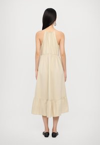 Beige puuvillasta valmistettu midi-mekko, jossa on kerätty vyötärö, naruolkaimet ja röyhelöhelma. Mallilla on päällään mustat liukuvartiset kengät.
