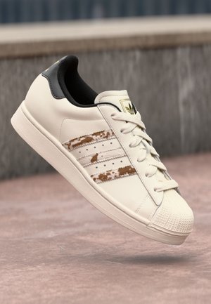 Biała tenisówka Adidas z czarnym obcasem, z wycieranymi brązowymi paskami i białymi sznurówkami, zawieszona nad różową powierzchnią.
