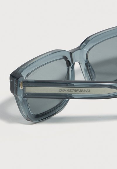 Κοντινό πλάνο ημιδιάφανων γκρι γυαλιών ηλίου με σκουρόχρωμους φακούς και λογότυπο Emporio Armani στο μπράτσο, σε απλό λευκό φόντο.