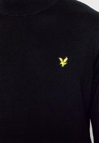 Pull à col roulé noir avec une texture côtelée, présentant un petit logo aigle brodé jaune sur le côté gauche de la poitrine.