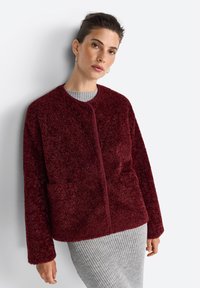 Une femme aux cheveux courts porte une veste en polaire bordeaux sur un pull gris à côtes, posant contre un fond clair.
