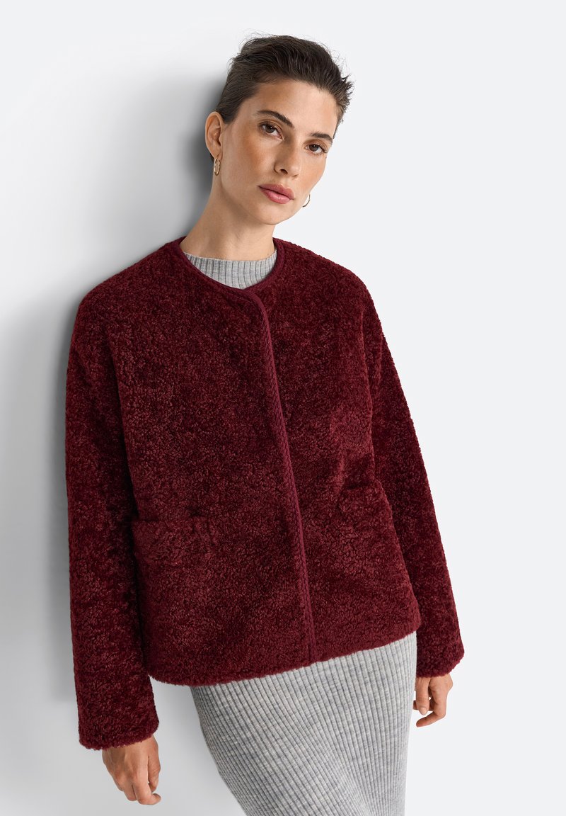 Une femme aux cheveux courts porte une veste en polaire bordeaux sur un pull gris à côtes, posant contre un fond clair.