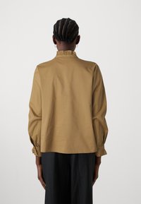 Blouse en coton marron avec un col froncé, des manches longues et des poignets froncés. La vue arrière montre un design lisse et minimaliste.