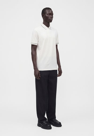 Homme debout face à l’avant, portant un polo blanc à manches courtes, un pantalon noir et des chaussures noires sur un fond uni clair.