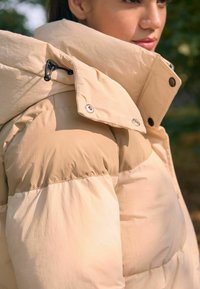 Beige dunjakke med høj krave, der har en blanding af matte og glansfulde teksturer, knap- og snoredetaljer samt quiltede sektioner.