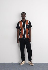 Camisa negra de manga corta con rayas verticales en naranja y blanco, bolsillo en el pecho, combinada con pantalones negros y zapatillas en color beige-marrón.