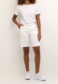 T-shirt en coton blanc associé à des shorts blancs arrivant au genou, dotés de poches latérales et de bords retroussés, complété par des baskets de sport blanches.
