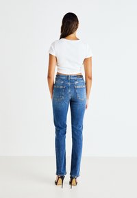 Τζιν παντελόνι με ψηλή μέση σε μπλε απόχρωση, με ίσια γραμμή, που διαθέτει πίσω τσέπες και ορατή ετικέτα στη μέση, συνδυασμένο με ένα λευκό κοντό μπλουζάκι.