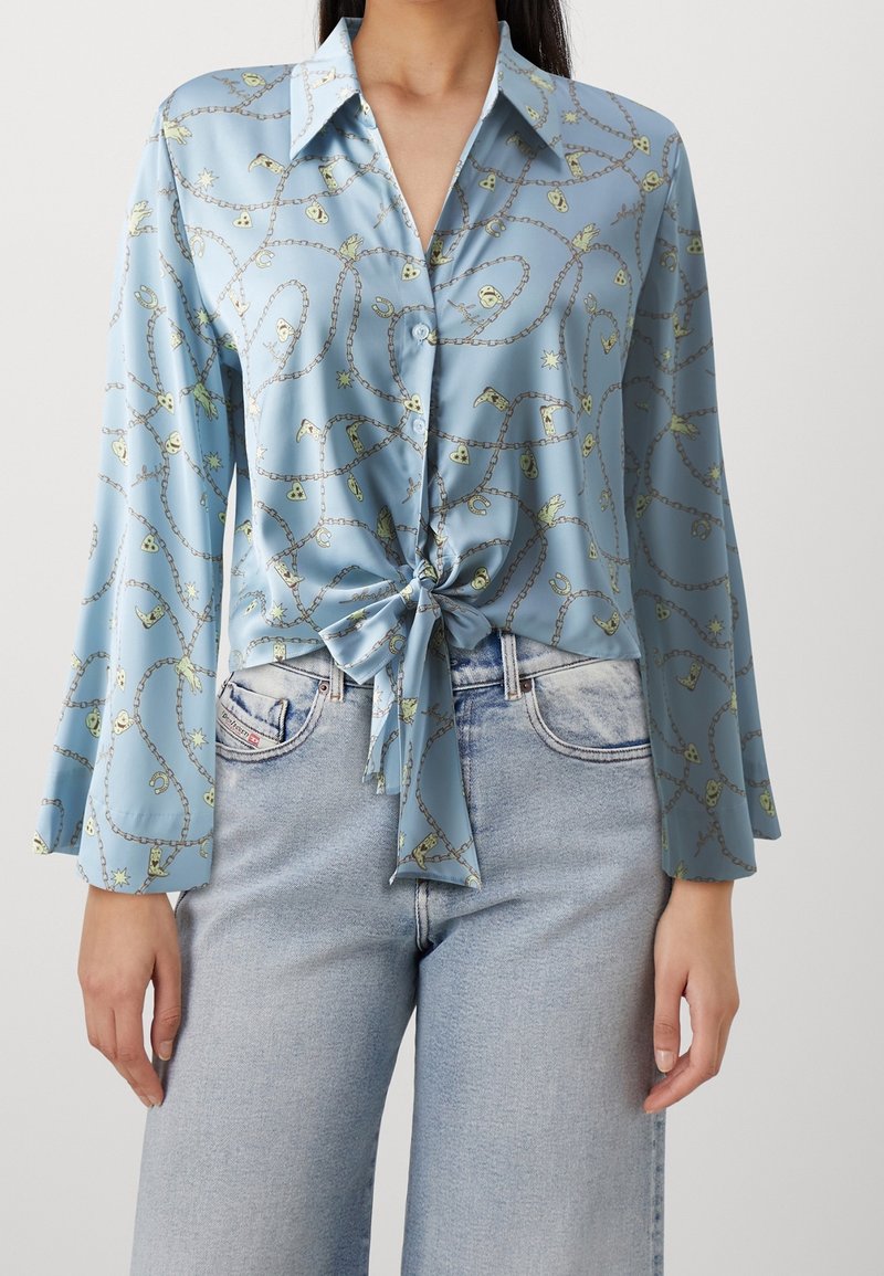 Femme portant un chemisier bleu clair boutonné avec un motif chaîne et breloques, noué devant, associé à un jean taille haute délavé clair.
