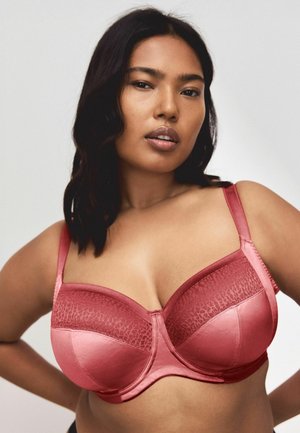 Woman with medium-length dark hair wearing a red satin and lace underwire bra, posing with hands on hips against plain background.