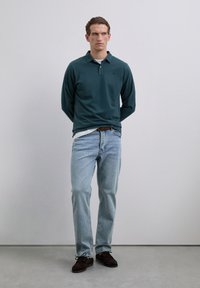 Joven de pie en interiores, vistiendo un polo de manga larga verde oscuro, jeans azul claro y zapatos marrones oscuros, mirando hacia un lado.