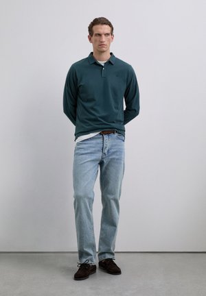 Joven de pie en interiores, vistiendo un polo de manga larga verde oscuro, jeans azul claro y zapatos marrones oscuros, mirando hacia un lado.