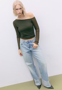Olivenfarvet off-the-shoulder langærmet top parret med lyseblå slidte bredbenede jeans og spidse sorte hæle, poseret mod en ensfarvet baggrund.