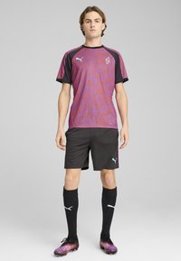 La camiseta de fútbol en morado y negro presenta un patrón de olas. Los pantalones cortos negros, los calcetines negros y los zapatos de fútbol coloridos completan el look deportivo.