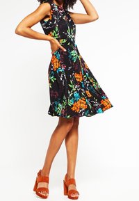 Femme portant une robe noire sans manches avec un motif floral éclatant orange, bleu et vert, et des sandales à talons hauts orange, posant avec la main sur la taille.