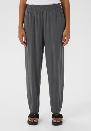 Graue Hose mit elastischem Bund, vertikalen Falten und lässiger Passform. Getragen mit schwarzen Sandalen, die ein geknotetes Riemendesign haben.