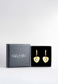 Guld hjärtformade örhängen med klara ädelstenar, visade i en svart box med ett präglat logotyp.