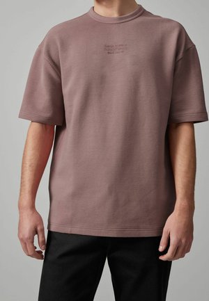 Homme portant un t-shirt mauve côtelé à manches courtes avec un petit texte embossé sur la poitrine, associé à un pantalon noir.