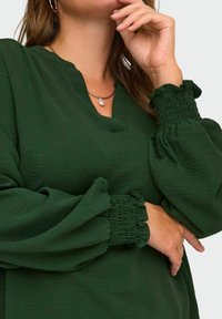 Donna che indossa una blusa verde scuro con maniche a sbuffo texture e polsini arricciati, accessoriata con una collana d'argento che tiene un pendente di perla.