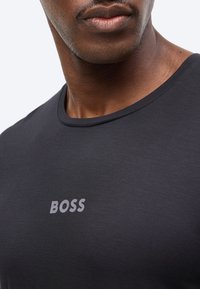 Κοντινό πλάνο ενός ατόμου που φοράει μαύρο μπλουζάκι με την επιγραφή «BOSS» σε γκρι χρώμα στο στήθος.