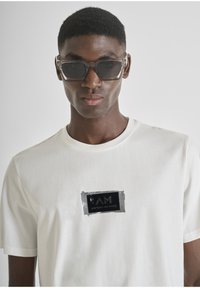 Camiseta blanca de algodón con un logo cuadrado negro que presenta el texto "AM" y "ANTONY MORATO". Combinada con gafas de sol transparentes y angulares.