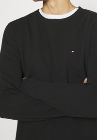 Schwarzes Langarmshirt aus strukturiertem Stoff mit einem weißen gerippten Kragen und einem kleinen, roten und weißen, gestickten Logo auf der Brust.
