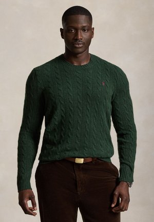 Uomo che indossa un maglione lavorato a trecce verde scuro e pantaloni marroni con una cintura marrone su uno sfondo beige uniforme.