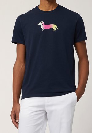 Mężczyzna ubrany w granatowy t-shirt z kolorowym gradientowym nadrukiem jamnika na piersi oraz białe spodnie, stojący z jedną ręką w kieszeni.