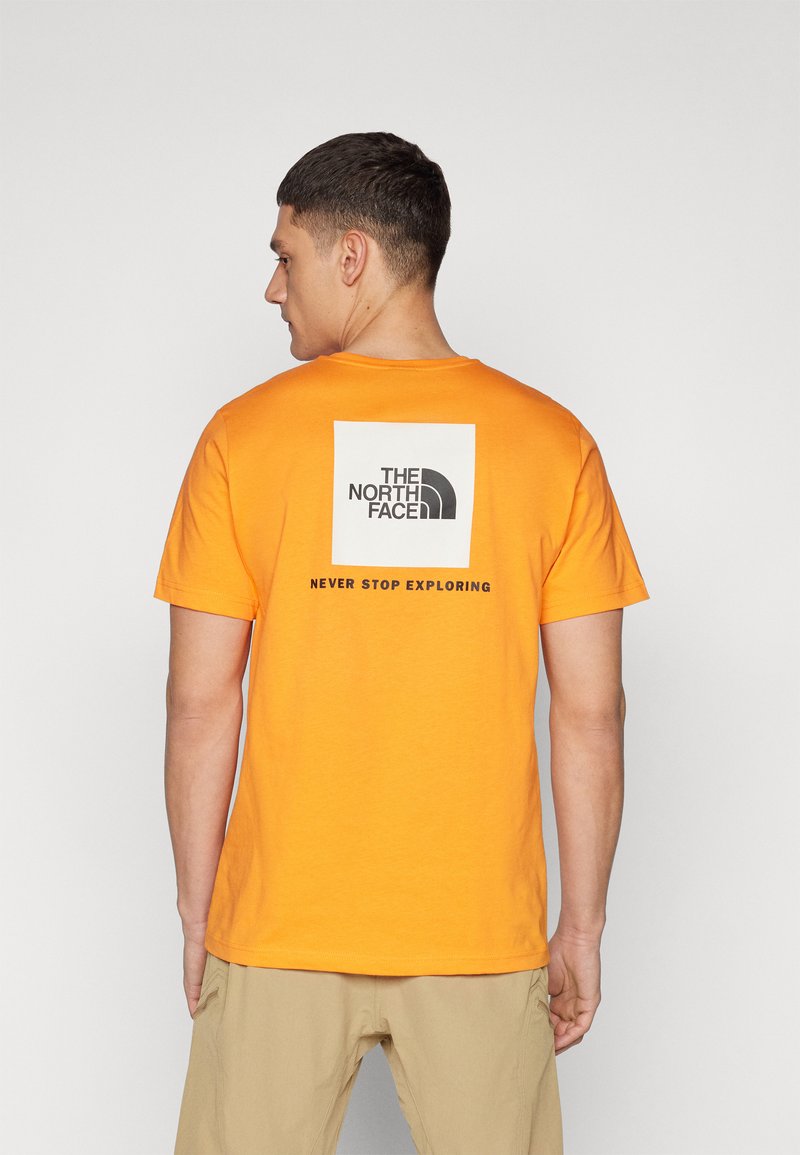 Oranžinė medvilninė marškinė su dideliu juodu kvadratiniu logotipu, kuriame užrašyta "THE NORTH FACE" ir "NEVER STOP EXPLORING" žemiau. Trumpomis rankovėmis.