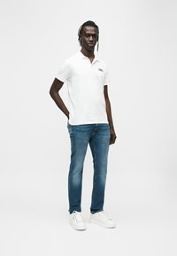 Λευκό πόλο πουκάμισο με γιακά και λογότυπο "KARL", σε συνδυασμό με μπλε τζιν και λευκά αθλητικά παπούτσια. Το μοντέλο στέκεται μπροστά σε απλό φόντο.