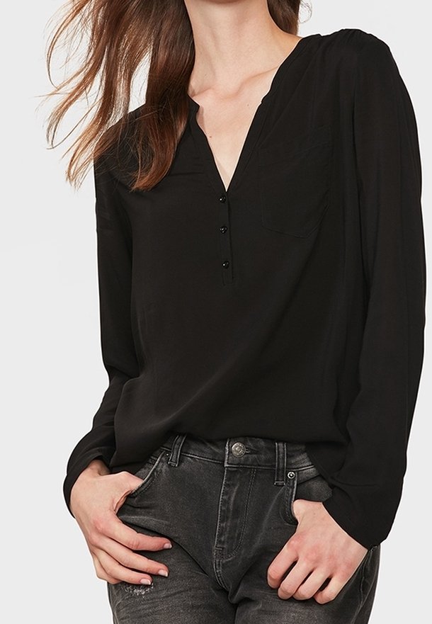 Donna che indossa una blusa nera a maniche lunghe e morbida con scollo a V e bottoni, abbinata a jeans grigi scuri, con le mani in tasca.