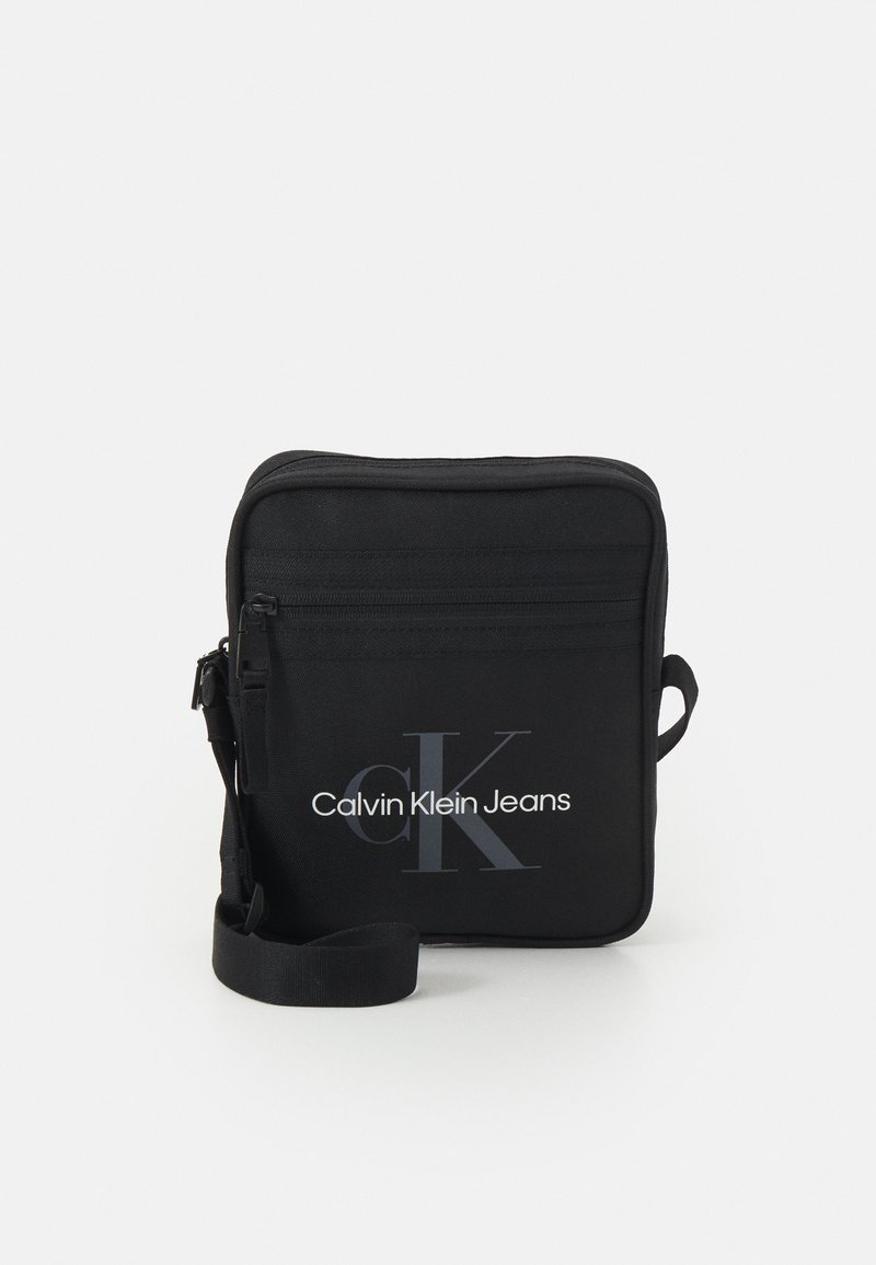 Bandolera rectangular negra con un bolsillo frontal con cierre. Cuenta con una correa para el hombro y el logo "Calvin Klein Jeans" en blanco.