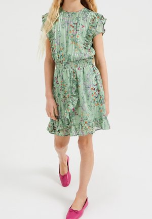 Ung person iført en grøn blomster-mønstret kjole med flæser og elastisk talje samt lyserøde ballet flats, stående mod en ensfarvet baggrund.