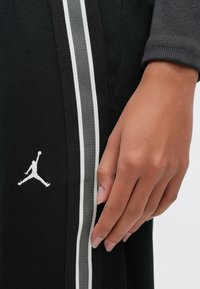 Hand rustend naast zwarte sportbroek met witte en grijze strepen en Jumpman-logo in close-up.