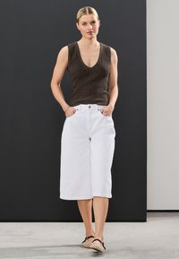 Braunes ärmelloses Oberteil mit strukturiertem Stoff, kombiniert mit lockeren weißen Culottes und hellem Schuhwerk. Einfaches Design vor neutralem Hintergrund.