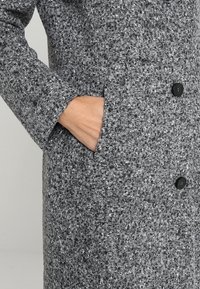 Šedý kabát se strukturou, zdobený tkaným vzorem, s knoflíkovým zapínáním a bočními kapsami. Materiál působí teplým a mírně lesklým dojmem.